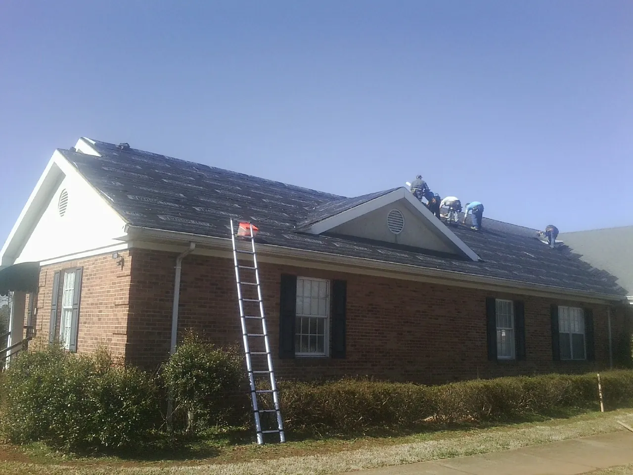Crew tearing shingles off a brick ranch roof with a ladder set — Emergency Roof Repair work by our crew in Minot