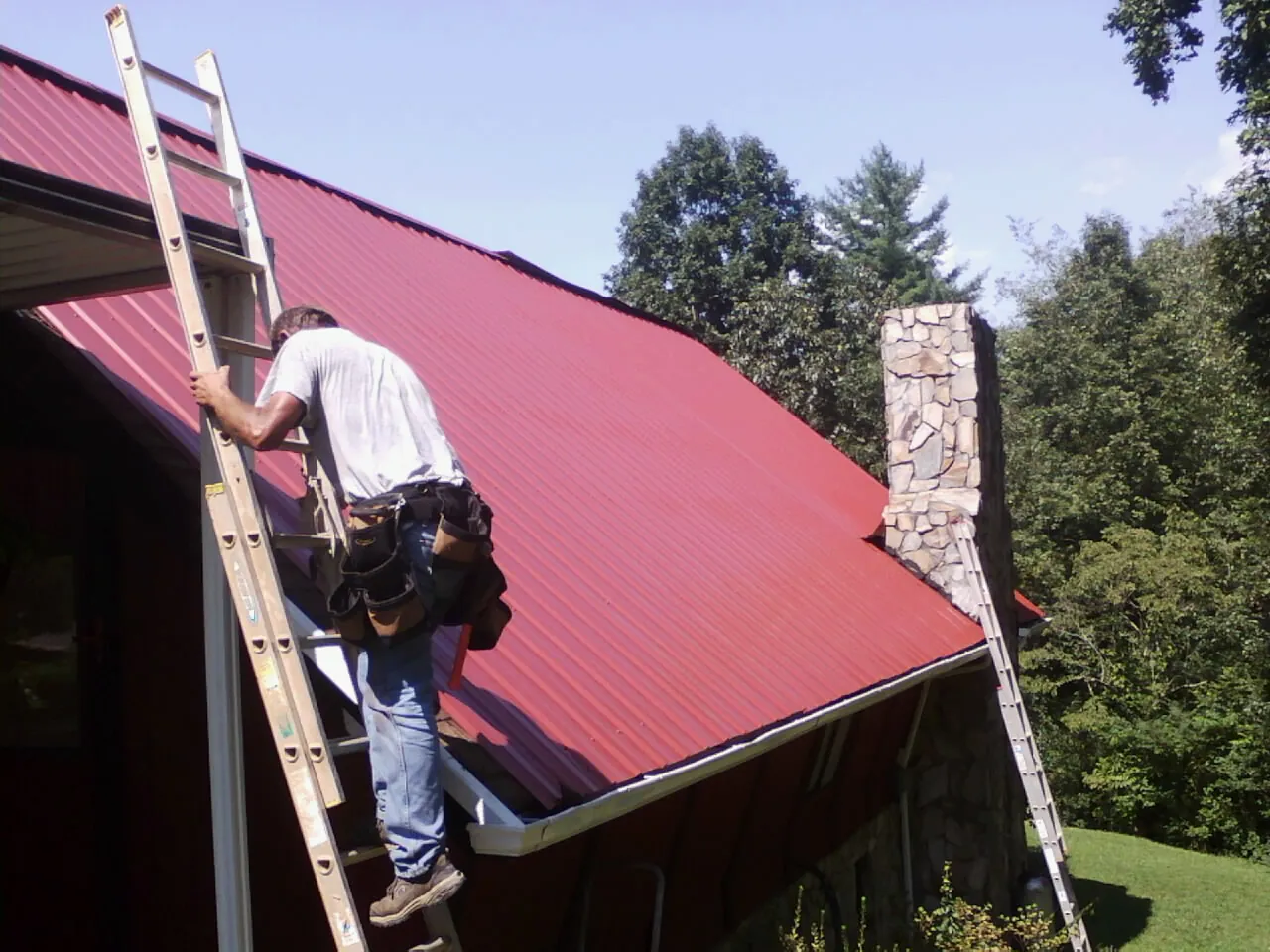 Roofer on a ladder beside a red metal roof and stone chimney — Soffit & Fascia Repair by Roofing Brothers in Minot